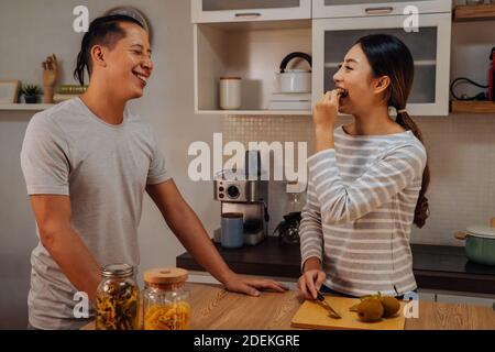 Young married couple cooking together in kitchen at home. Young woman feeding her man with kiwi and fruits indoors Stock Photo