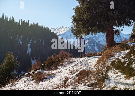 A bearded male traveler in sun glasses stands on a mountain trail in winter. Trekking outdoors in the mountains in winter on a nice sunny day Stock Photo