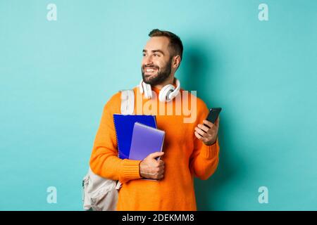 Education. Handsome male student with headphones and backpack, using mobile phone and holding notebooks, smiling happy, standing over turquoise Stock Photo