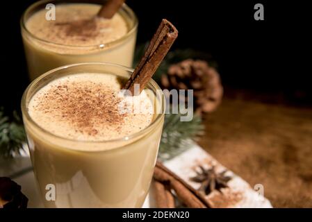 Homemade traditional Christmas eggnog drinks in glasses with ground nutmeg, cinnamon and decorating items on wood table, preparing for celebrating fes Stock Photo