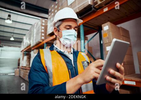 Male factory worker using digital tablet wearing protective mask at warehouse Stock Photo