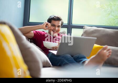 YOUNG MAN EATING CHIPS FROM A BOWL IN HIS HAND WHILE WATCHING A MOVIE ON HIS LAPTOP Stock Photo