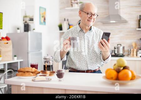 Portrait of elderly man smiling while using modern smartphone in the kitchen. Internet online chat technology video webcam making a video call connection camera communication conference call Stock Photo