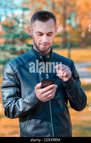 Youthful Guy Using Black Smartphone at the Beautiful Autumn Park. Handsome Young Man with Mobile Phone at Sunny Day - Medium Shot Portrait Stock Photo