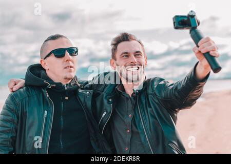 Two Handsome Smiling Friends Making Selfie Using Action Camera with Gimbal Stabilizer at the Beach. Youthful Men in Black Clothes Having Fun by Making Photos Stock Photo