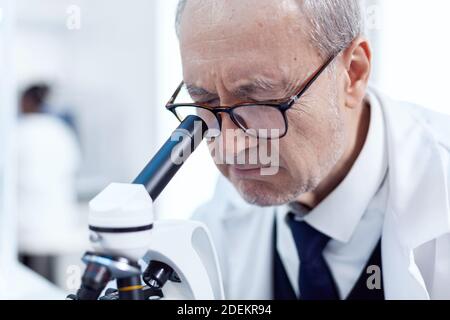 Close up of senior scientist analyzing virus sample using microscope in modern facility. Chemist researcher in sterile lab doing experiments for medical industry using modern technology. Stock Photo