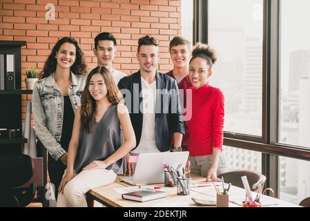 Young trendy diverse smiling millennial team in casual clothes around office table after business meeting with red brick wall and window city skyline Stock Photo