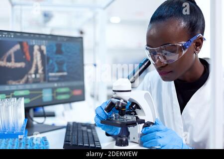 African pharmacology researcher analysing chemical substance using microscope for study.. Black healthcare scientist in biochemistry laboratory wearing sterile equipment. Stock Photo