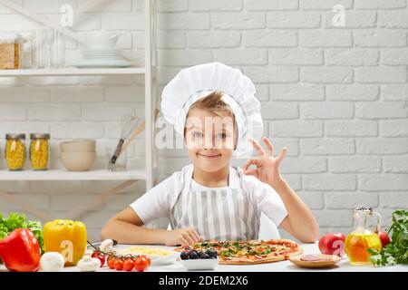 little boy in chef hat and an apron cooking pizza in the kitchen. child showing Ok gesture. delicious Stock Photo