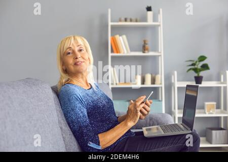 Mature woman sitting on couch with laptop, holding her cell phone and looking away Stock Photo
