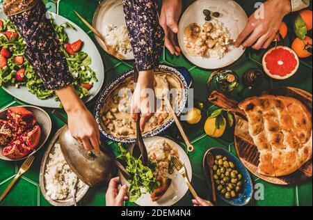 Family having Turkish dinner. Flat-lay of people hands taking lamb in yogurt sauce, arugula and strawberry salad, rice pilav and flatbread over green table, top view. Gathering, Ramazan iftar supper Stock Photo