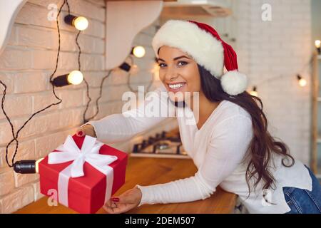 Smiling woman in Santa hat holding Christmas present leaning over kitchen countertop Stock Photo