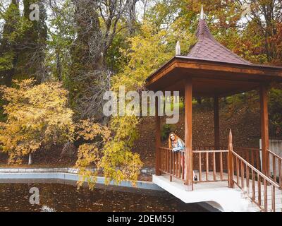 Redhead girl stands in a gazebo over muddy water in an autumn park Stock Photo