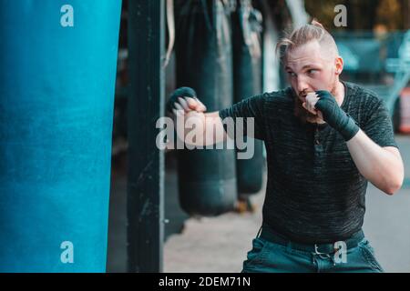 Street Fighter in Black Clothes and Bandages on the Wrist Boxing in Punching Bag Outdoors. Young Man Doing Box Training and Practicing His Punches at the Outside Gym Stock Photo