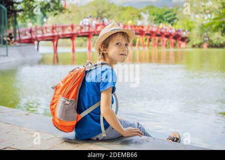 Caucasian boy tourist on background of Red Bridge in public park garden with trees and reflection in the middle of Hoan Kiem Lake in Downtown Hanoi Stock Photo