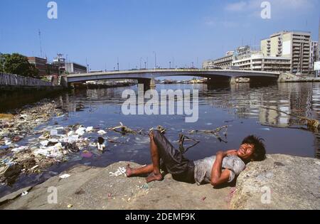 A poverty-stricken homeless man beside the Pasig River in Manila, Luzon Island, the Philippines. The homeless man amassed whatever items he might need from the trash that floated past him on the river. The Jones Bridge, linking Ermita to Binondo (Manila's Chinatown), is seen in the background. Stock Photo