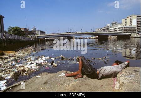 A poverty-stricken homeless man beside the Pasig River in Manila, Luzon Island, the Philippines. The homeless man amassed whatever items he might need from the trash that floated past him on the river. The Jones Bridge, linking Ermita to Binondo (Manila's Chinatown), is seen in the background. Stock Photo