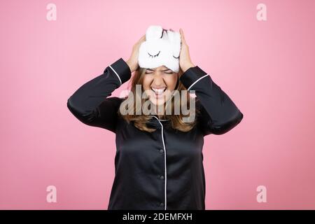 Young caucasian woman wearing sleep mask and pajamas over isolated pink background suffering from headache desperate and stressed because pain and mig