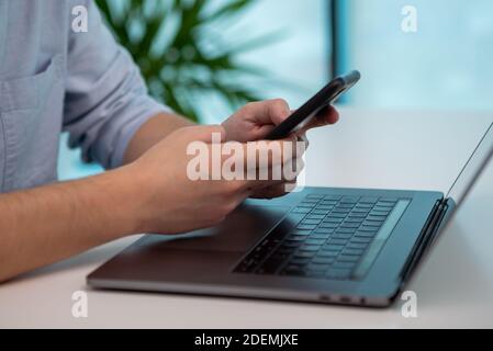 Close up of a man's hands typing on his smartphone in the office. Then he puts down the mobile phone and starts typing on the laptop. Stock Photo