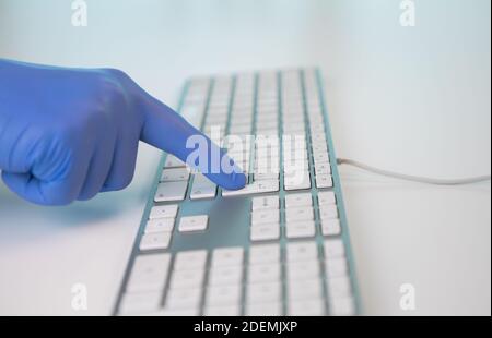 Gloved hand of a healthcare worker typing on a computer keyboard. Pressing the enter key. Consultation of a hospital where Coronavirus or Covid 19 is Stock Photo