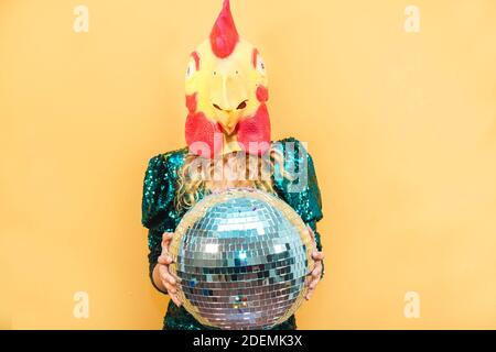 Young woman wearing chicken mask while holding disco ball at new year's party - Soft focus on face Stock Photo