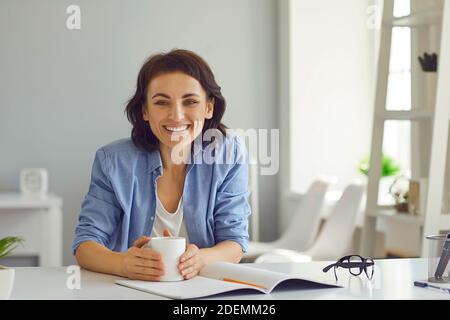 Happy woman sitting at desk with open notebook and cup of coffee smiling and looking at camera Stock Photo