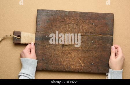 two female hands hold an empty old brown rectangular wooden cutting board, brown background, top view Stock Photo