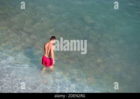 Man in red shorts going to swim in a sea. Aerial view to the pebble beach with transparent water, background for vacation and travel Stock Photo