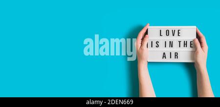 Festive banner with woman hands holding glowing lightbox with text Love is in the Air on bright blue background for Valentine's Day. Flatlay banner fo Stock Photo