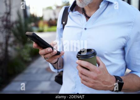 In touch. Close up shot of hands of modern middle aged businessman using his smartphone while walking outdoors with a cup of coffee Stock Photo