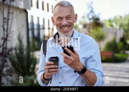 Happy modern middle aged businessman smiling while using his smartphone, walking outdoors with a cup of coffee Stock Photo