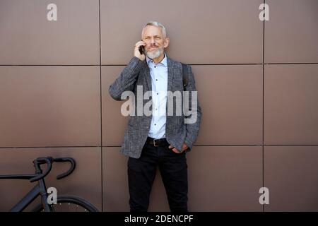 Modern middle aged businessman looking thoughtful while talking on the phone standing with his bicycle outdoors Stock Photo