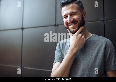Portrait of a beautiful stylish guy, hipster dressed in a gray empty t-shirt standing on a black wall background. Stock Photo