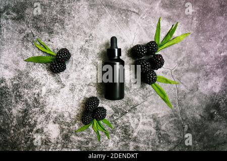 Black bottle of men's facial anti-aging serum on a dark grunge cement background surrounded by blackberries rich in antioxidants. Stock Photo