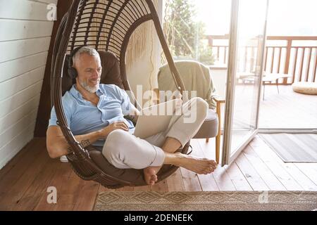 Happy middle aged caucasian businessman wearing headphones using laptop while sitting on swing chair at his modern apartment and working from home Stock Photo