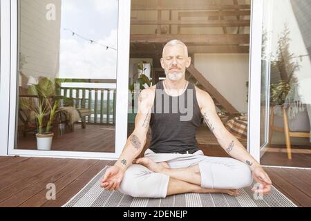 Balance in life. Relaxed middle aged caucasian man sitting in lotus pose on the sunny terrace of his modern flat and meditating or practicing yoga Stock Photo