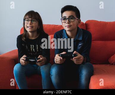 Two siblings, a boy and a girl, playing the game console sitting on the couch at home. Stock Photo