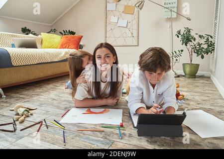 Family time. Teenage boy lying on the floor in living room, using digital tablet and doing homework online while his mother and sister playing Stock Photo