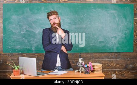 Teacher interesting interlocutor best friend. Teacher charismatic hipster stand near table classroom chalkboard background. Talking to students or pupils. Teacher bearded man tell interesting story. Stock Photo