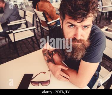Guy having rest at cafe terrace. Hipster, client, visitor on serious face sits at cafe outdoor. Man with beard and mustache in calm mood while relaxing at cafe terrace. Bearded macho concept. Stock Photo
