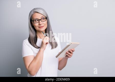 Photo portrait of smart old businesswoman writing notes in notebook looking at blank space isolated on grey color background Stock Photo