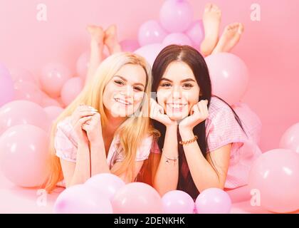Gossip concept. Blonde and brunette on smiling faces have fun at domestic bedroom party. Girls lay on belly near balloons, pink background. Sisters or best friends in pajamas at girlish pajamas party. Stock Photo