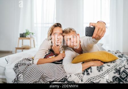 Cheerful young adults сouple in pajamas taking a Funny grimace selfie photo using a modern smartphone as they lazy relaxing lying in a cozy bed in the Stock Photo