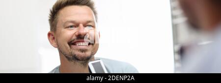 Smiling man stands in front of a mirror and holds shaving razor in his hands. Stock Photo