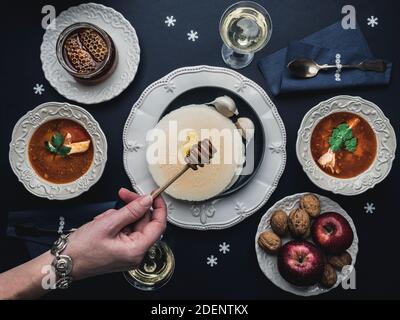 Female hand pouring honey on Christmas Eve wafers. Table with fish soup, wafers, honey, apples, walnuts, white wine and decoration. Overhead view. Stock Photo