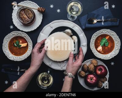 Female hands holding Christmas Eve wafers. Table with fish soup, wafers, honey, glasses of white wine and decoration. Overhead view. Stock Photo