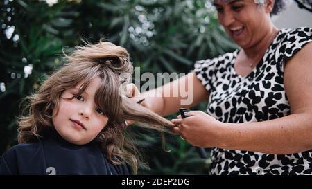 Stock photo of young girl getting his haircut by a happy woman in the garden. She is happy and looking at camera. Stock Photo