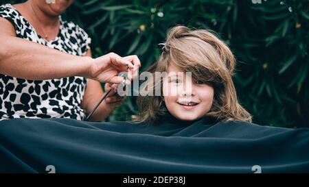 young girl getting his haircut by a woman in the garden. She is happy and looking at camera. Stock Photo