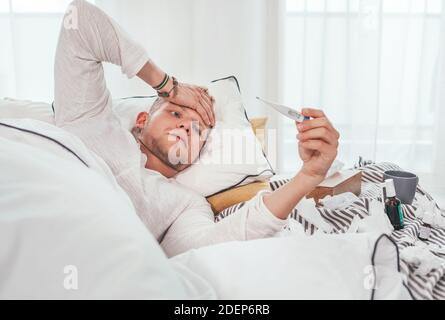 Tired and unhealthy looking at a medical thermometer as lying in a cozy home bed beside a lot of used paper tissues and medicines on the blanket. Seas Stock Photo