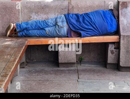 Homeless man sleeping rough on a wooden park bench during the day. A symbol of the growing problems of homelessness and poverty especially in the citi Stock Photo